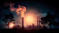 Industrial factory silhouette at sunset with smoke stacks emitting steam against orange sky and dark trees framing dramatic atmospheric scene.