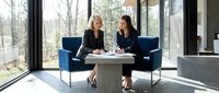 Two professional businesswomen having meeting in modern office lounge with blue chairs and large windows overlooking nature for corporate collaboration.