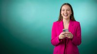 Young professional woman in bright pink blazer laughing while holding smartphone against turquoise background for business and technology concepts.