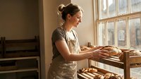 Female baker with medium complexion arranging fresh baked bread loaves on wooden shelves near sunlit window in artisan bakery shop or home kitchen setting.