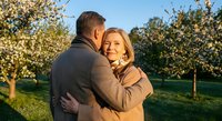 Happy mature couple embracing in blooming spring orchard during golden hour sunset. Middle-aged man and woman sharing romantic moment among flowering trees.