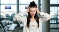 Stressed businesswoman with dark hair and fair complexion holding her head in frustration inside modern office with large windows and city view, expressing work-related stress and headache.