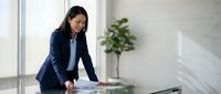Professional Asian businesswoman in navy suit reviewing documents at modern office desk with natural lighting and plants in background for corporate presentations.