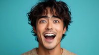 Young Asian male expressing extreme excitement and joy with wide open eyes and mouth against turquoise background, showing genuine surprise and happiness.