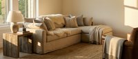 Modern beige sectional sofa with throw pillows and blanket in bright living room with natural sunlight streaming through large windows creating warm atmosphere.