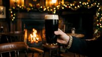 Hand holding a pint glass of dark beer in front of a cozy fireplace with warm golden bokeh lights and holiday garland decorations in background.