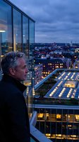 Middle-aged Caucasian male professional gazing at illuminated cityscape from high-rise balcony at dusk. Urban business concept for corporate success and leadership themes.