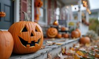 Carved jack o'lantern pumpkin with classic triangle eyes and smile on front porch steps decorated for Halloween, with autumn leaves and glowing lights in background.