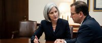 Senior woman with gray hair and man in business suits discussing at wooden conference table in office setting.
