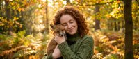 Middle-aged Caucasian woman with curly red hair embracing small tabby kitten in autumn forest. Warm golden sunlight filters through colorful fall foliage.