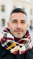 Middle-aged Hispanic man with salt and pepper hair smiling warmly at camera, wearing plaid scarf in red, black and yellow. Soft natural lighting creates welcoming expression.