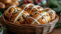 Fresh hot cross buns with raisins and glaze in woven basket, traditional Easter holiday baking, close up view on rustic wooden background with greenery.