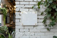 Blank white square signboard mockup on rustic white brick wall with green climbing plants and decorative foliage, perfect for branding and outdoor business display design.
