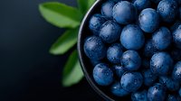 Fresh ripe blueberries with water drops in dark ceramic bowl on black background with green leaves, macro food photography for healthy eating.