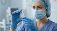 Female healthcare worker in surgical mask and cap holding medical vial in laboratory. Professional nurse or doctor preparing medication in clinical setting.