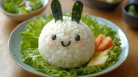 Cute bunny-shaped rice ball with cucumber ears and nori face details served on lettuce with fresh vegetable garnish on blue ceramic plate, creative food styling.