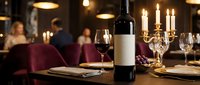 Elegant restaurant table setting with wine bottle and glasses in upscale dining room with warm ambient lighting and blurred diners in background.