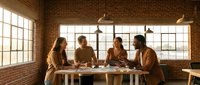 Diverse group of four young professionals gathered around wooden table in modern loft office with brick walls and natural light from large windows, collaborating on project.