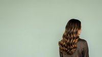 Young woman with long wavy brunette hair and fair complexion wearing dark shirt, standing against neutral green background, rear view for portrait and lifestyle use.