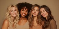 Four diverse women with different hair textures and skin tones smiling together against warm beige background, showcasing natural beauty and friendship.