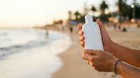 Person holding blank white sunscreen bottle at beach during golden hour sunset, displaying product for skincare and sun protection advertising.