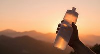 Hand holding sports water bottle against mountain sunset backdrop for hydration and outdoor fitness activities.