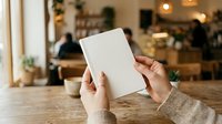 Person with medium skin tone holding blank white card in warm cafe setting with blurred background, suitable for marketing mockups and business presentations.