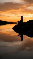 Silhouetted woman meditating on rocky riverbank at golden sunset, reflected in calm water below, conveying peace, mindfulness and tranquility in nature.