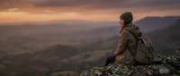 Male hiker wearing brown jacket and backpack sitting on rocky mountain peak at sunset, overlooking vast valley landscape below during golden hour adventure travel.