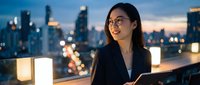 Young Asian businesswoman with glasses on urban rooftop terrace at dusk with city skyline and glowing lanterns creating professional evening atmosphere.