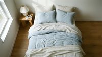 Overhead view of neatly arranged bed with light blue and cream bedding, wooden nightstand with lamp, and natural window light in modern bedroom interior for home decor and lifestyle content.