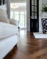 Modern bright living room with white sofa, black door, and herringbone hardwood flooring. Crystal chandelier and floral accent chair create elegant contrast.
