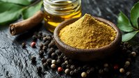 Ground curry powder in wooden bowl with spices, black peppercorns, cinnamon stick, honey jar and fresh green leaves on dark stone background.