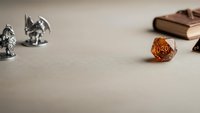Tabletop role-playing game setup with metal miniature figures, orange twenty-sided die, and leather-bound rulebook on neutral surface for gaming adventures.