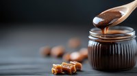Wooden spoon drizzling rich caramel sauce into glass jar with golden candies and nuts on dark background for dessert recipes and confectionery projects.