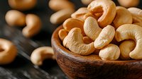 Fresh roasted cashew nuts in wooden bowl with scattered nuts on dark background, macro food photography showing natural texture and warm tones.