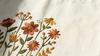Delicate hand embroidered wildflowers in warm autumn colors on natural linen fabric showcasing traditional needlework craftsmanship for home decor projects.