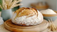 Freshly baked crusty sourdough bread on wooden cutting board with wheat ears and flour in background, rustic artisanal baking composition.