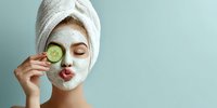 Young woman with white facial mask and towel on head holding cucumber slice over eye for spa skincare treatment and beauty routine.