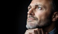 Thoughtful middle-aged Caucasian man with gray beard looking up contemplatively against dark background, hand on chin in reflective pose. Professional portrait.
