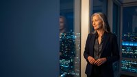 Professional blonde businesswoman in black suit standing by office window overlooking illuminated city skyline at night for corporate presentations.