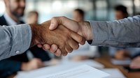 Close-up of diverse business professionals shaking hands over signed contract documents, with blurred colleagues in background at modern office meeting.