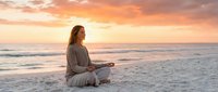 Young Caucasian woman meditating in lotus pose on sandy beach at golden sunset, practicing mindfulness and yoga for wellness and inner peace.