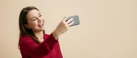 Happy young woman taking selfie with smartphone against beige background. Casual portrait of smiling female using mobile phone for social media content.