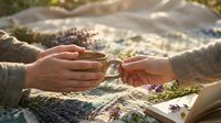 Two hands gently passing wooden spoon over blanket with dried lavender flowers during outdoor picnic in natural sunlight setting.