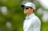 Young Caucasian male athlete in white polo shirt and baseball cap wearing sunglasses looking confident against blurred green background during outdoor sports event.