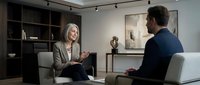 Professional blonde businesswoman in beige suit conducting interview or consultation with male client in modern office setting with contemporary decor.