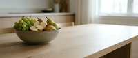 A wooden bowl filled with fresh green grapes, apples, and pears sits on a light natural wood table in a bright kitchen with soft window light.