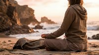 Woman meditating in lotus pose on sandy beach at golden hour sunset with rocky coastline and ocean waves creating peaceful mindfulness wellness scene.