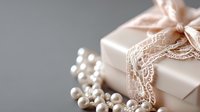 Elegant gift box wrapped in pink paper with delicate lace ribbon and scattered white pearls on gray background, romantic wedding or anniversary composition.